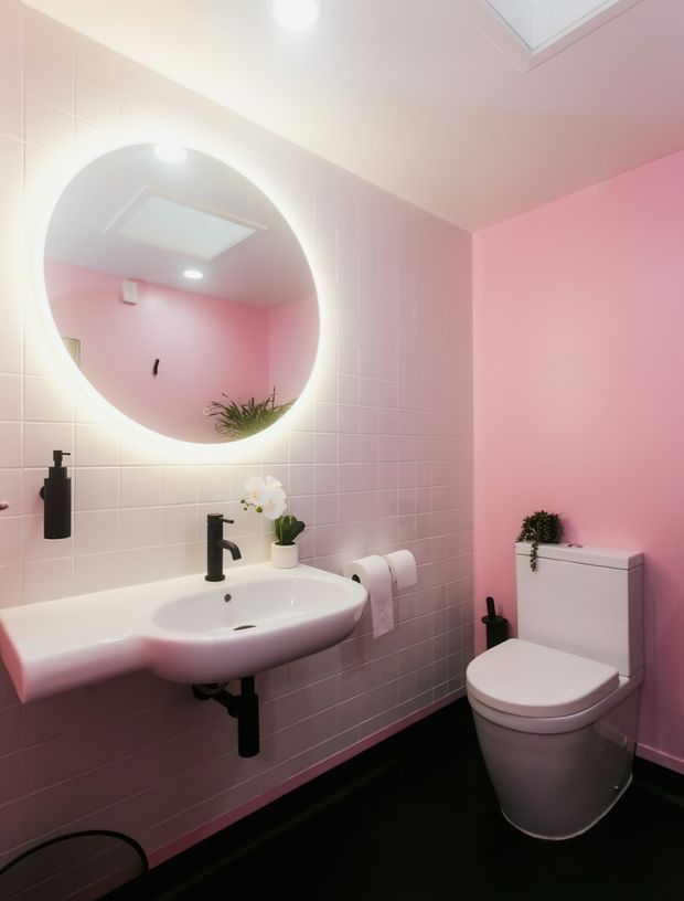 Compact contemporary bathroom with pink tiled walls, a large round backlit mirror, white wall-mounted basin, matte black tapware and accessories, and a white toilet. A skylight and recessed lighting brighten the space, while a small orchid and plant add softness.