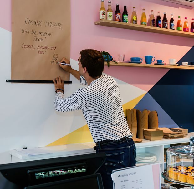 A colorful bakery or café kitchen area with a person writing on a large chalkboard-style wall panel. Open shelves display bottled drinks, cups, and small kitchenware, while a countertop holds bread, bowls, plates, and a covered cake display. The space features playful geometric wall shapes in pink, yellow, white, and dark blue, with bright, informal commercial interiors.