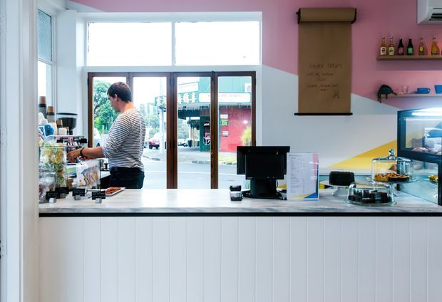 Bright, compact café or takeaway shop interior viewed from the customer side of the service counter. The foreground features a white vertically panelled counter with a dark benchtop, point-of-sale terminal, display case, cakes and drinks. Behind it, a staff member works near the front glazing and entrance. The walls use playful geometric colour blocking in pink, white, and yellow, with open shelving and a wall-mounted scroll or sign.