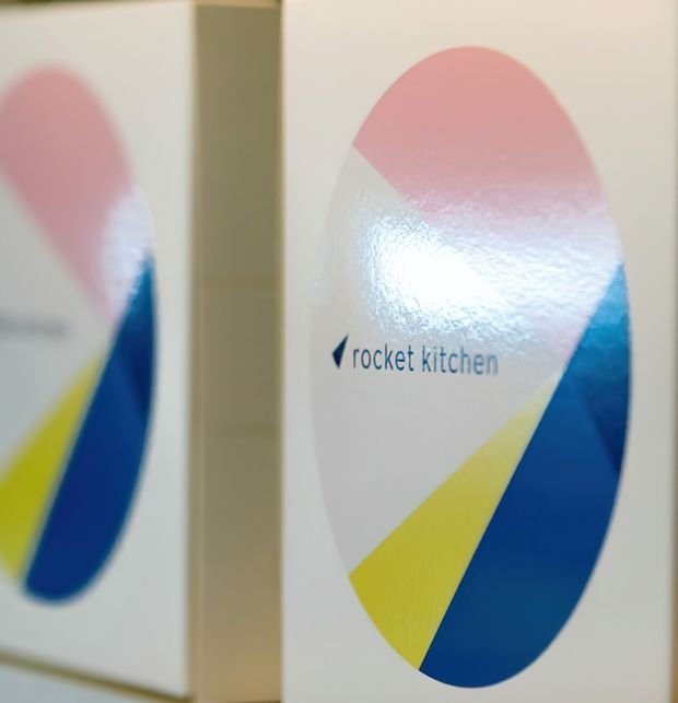 Close-up of a branded white panel or sign featuring the 'rocket kitchen' logo and a bold abstract oval graphic in pink, blue, and yellow. A partial mirrored or adjacent panel is visible on the left, with shallow depth of field and soft indoor lighting.