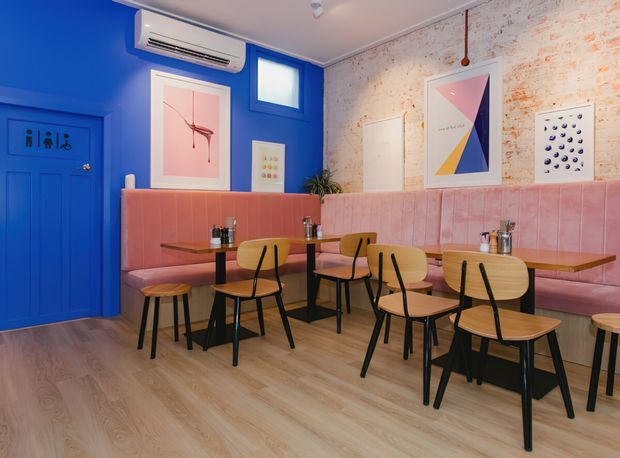 A colourful contemporary café or casual dining interior with vivid cobalt-blue walls and door, a long blush-pink upholstered banquette, timber tables, and black-framed wooden chairs and stools. Exposed white-painted brick forms the feature wall, with framed graphic artwork, pendant-free ceiling lighting, and a wall-mounted air-conditioning unit. The space has a warm timber floor and playful, high-contrast styling.
