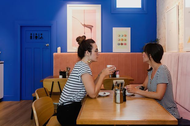 A brightly coloured café or restaurant dining area with vivid cobalt-blue walls and door, a long timber dining table, mustard-toned chairs, and a blush-pink upholstered banquette. Two diners sit opposite each other, with framed artwork, pendant lighting, and a small high window adding visual detail. The space has a playful, retro-inspired fit-out with strong colour contrast.