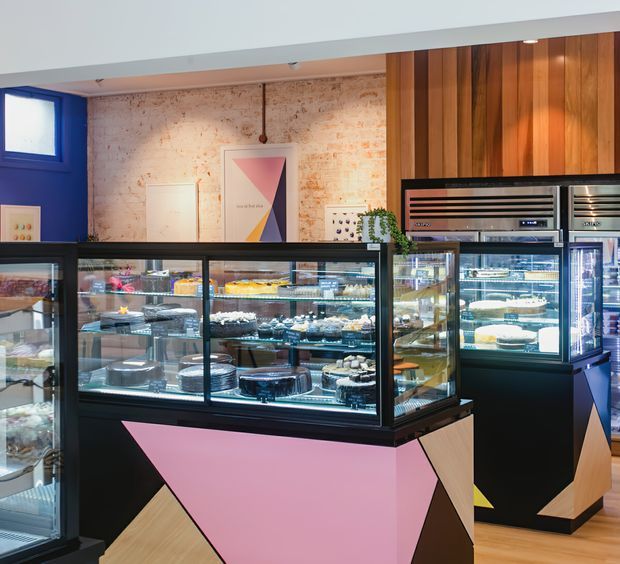 A brightly lit contemporary dessert or bakery retail interior with multiple black-framed glass display counters filled with cakes and pastries. The fit-out combines geometric black, pink, and cream counter panels, warm timber flooring and vertical timber wall cladding, exposed pale brick, and vivid blue wall accents. Pendant lighting and decorative artwork add colour to the commercial space.