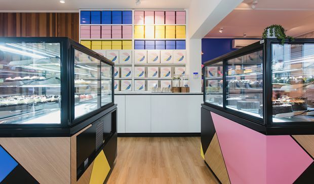 A brightly coloured contemporary dessert or bakery retail interior, viewed between two large glass display counters filled with cakes and pastries. The counters feature black frames and geometric panels in pink, blue, yellow and timber tones. A white service counter sits at the rear beneath a colourful grid artwork wall, with timber wall panelling and a small potted plant adding warmth.