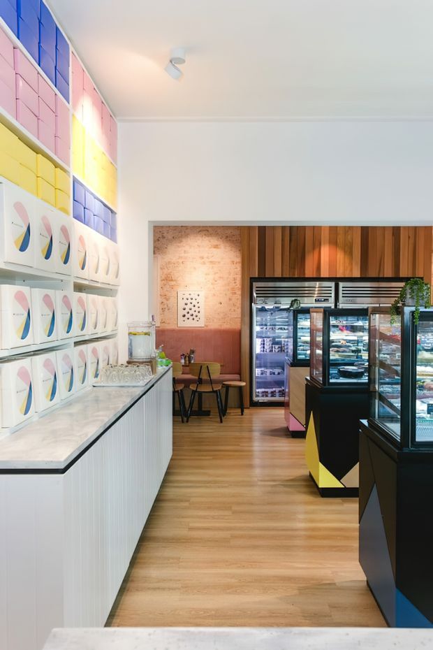 A bright, compact retail food-service interior with a long white fluted counter and pale stone benchtop on the left, colorful geometric product display shelving above it, and dark glass-front refrigerated display cases on the right. The rear wall combines exposed brick with vertical warm timber panelling, while light timber flooring and white walls create a clean, contemporary atmosphere. Small café-style seating is visible at the back.