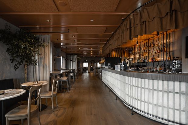 A long, atmospheric hospitality interior featuring a curved tiled bar with illuminated bottle shelving, dark timber flooring, timber-panelled ceilings, upholstered booth seating, and small round tables. The space has warm ambient lighting and a refined contemporary restaurant or cocktail-bar character.