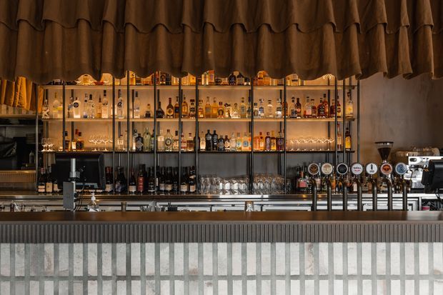 A stylish commercial bar counter viewed straight on, with illuminated glass shelving displaying liquor bottles and glassware, numerous beer taps, coffee equipment, and a textured tiled bar front. A dark, pleated fabric canopy hangs above, creating a warm, moody hospitality atmosphere.