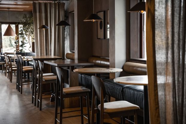 A moody, refined restaurant or hospitality dining interior with rows of dark timber tables, upholstered banquette seating, black-framed chairs and stools, pendant lamps, concrete-look walls, and full-height curtaining. Large windows and mirrors/reflections extend the view of the dining space.