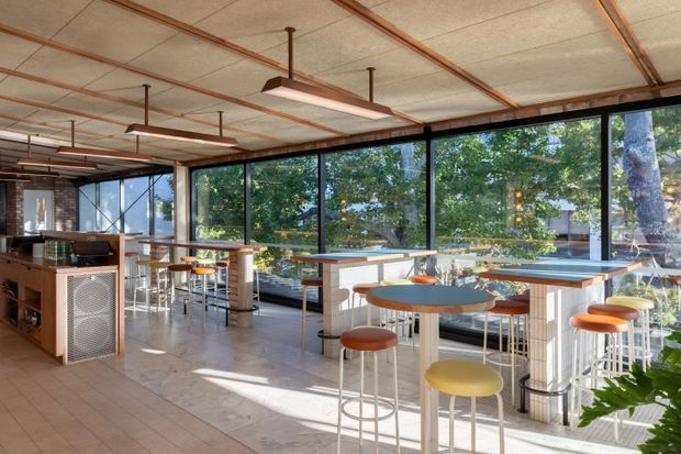 Bright contemporary commercial interior with a café or hospitality-style seating area, featuring floor-to-ceiling glazing, multiple high tables and colourful round stools, a service counter, and a tiled floor. Exposed copper-toned ceiling conduits and suspended rectangular light fixtures add an industrial character, while extensive windows provide views to leafy surroundings.