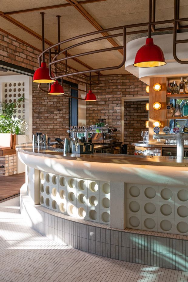 Interior view of a stylish bar or hospitality space featuring a curved white tiled bar counter with circular relief panels, exposed red brick walls, dark timber and metal ceiling structure, and multiple red pendant lights. Beer taps, bottles, shelving, and bar equipment are visible behind the counter, with strong natural sunlight creating patterned shadows across the floor and counter.
