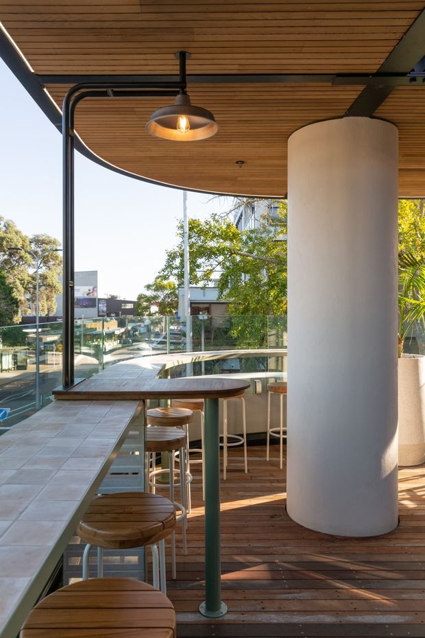 A bright elevated outdoor hospitality terrace with a curved timber-lined canopy ceiling, integrated warm pendant lighting, tiled bar counter, timber stools, glass balustrades, and a large cylindrical white column. The terrace overlooks a leafy urban streetscape and includes a curved built-in counter with seating.