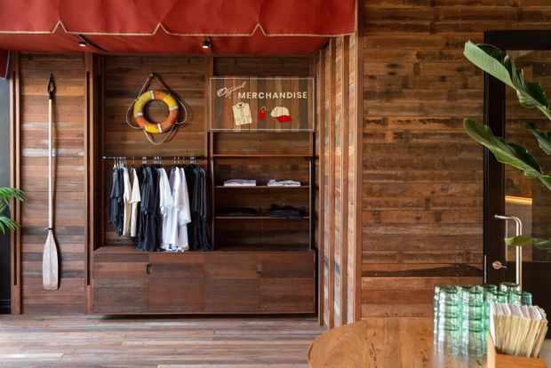 A warmly lit retail merchandise display with dark timber wall panelling and built-in cabinetry. Clothing hangs beside open timber shelving, with a framed merchandise sign, decorative nautical objects, and a red fabric awning overhead. A timber-framed glass door and a small counter or table with packaged goods are visible at right.