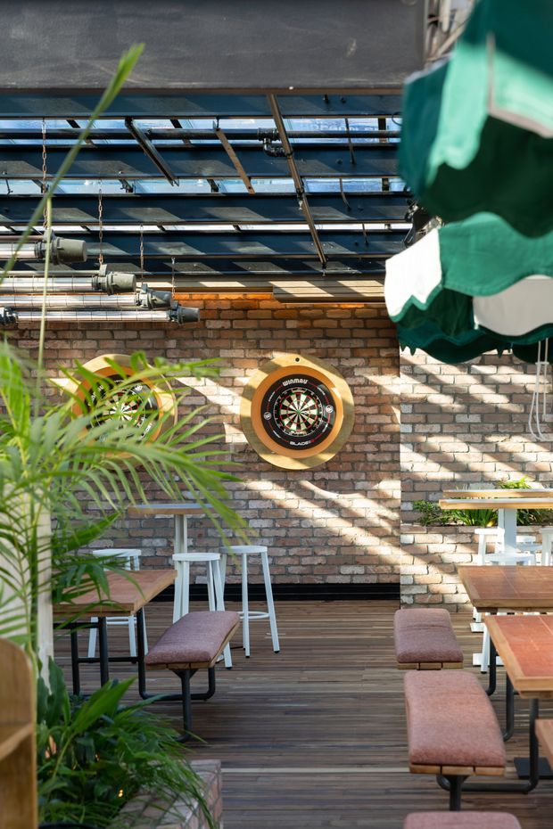 A bright industrial-style dining and social space with exposed brick walls, an open steel-and-glass roof structure, visible ductwork, timber flooring, and communal wooden tables with upholstered benches. Tropical plants, a dartboard, and strong sunlight create a relaxed hospitality or commercial venue atmosphere.