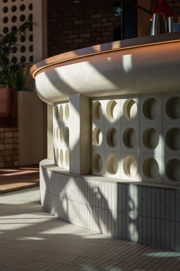 Close-up architectural photograph of a sculptural curved bar or reception counter. The counter features a rounded white plastered underside, a dark stone or metal countertop with warm integrated edge lighting, and a base clad in patterned breeze-block tiles with circular recesses. The surrounding space has tiled flooring, exposed dark brickwork, indoor planting, and dramatic sunlight and shadow patterns, creating a retro-modern hospitality interior.