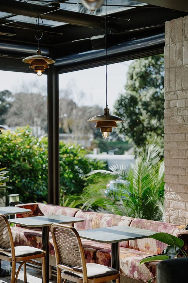 A stylish cafe or restaurant dining area with cane-back chairs, small square tables, and a long patterned upholstered banquette beside expansive black-framed glazing. Brass pendant lights hang from an exposed black ceiling with visible services, while a pale brick column and lush greenery outside add warmth and an indoor-outdoor atmosphere.