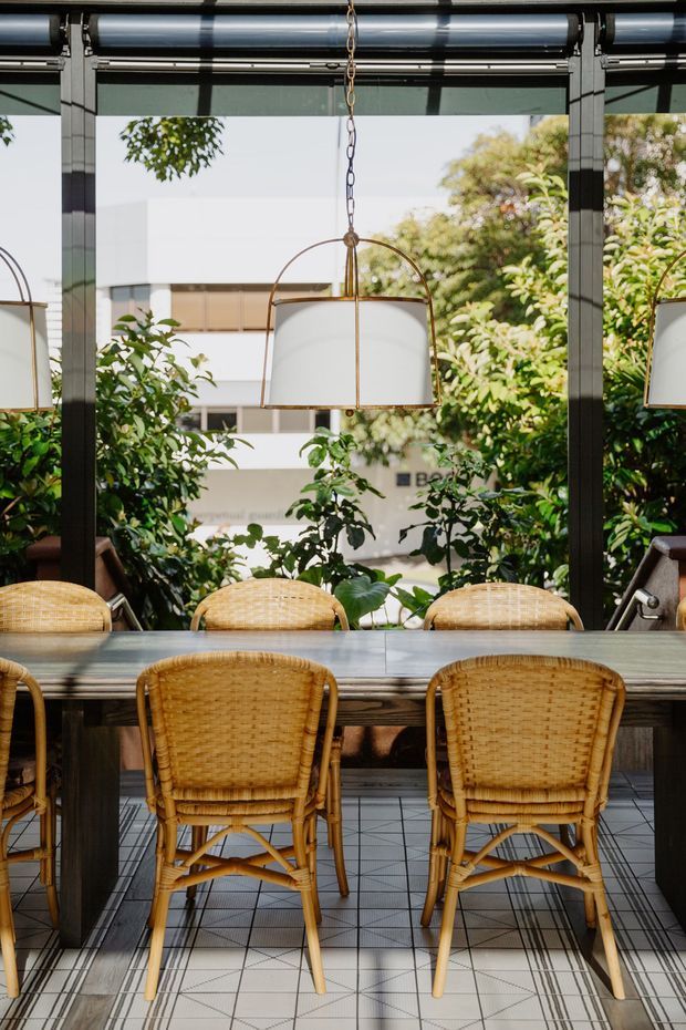 A refined dining area enclosed by dark-framed floor-to-ceiling glazing, with a long dark timber dining table, woven rattan chairs, and suspended drum pendant lights. Lush greenery is visible outside and reflected in the glazing, creating a light-filled indoor-outdoor atmosphere. The floor is finished with small square tiles in a geometric grid.