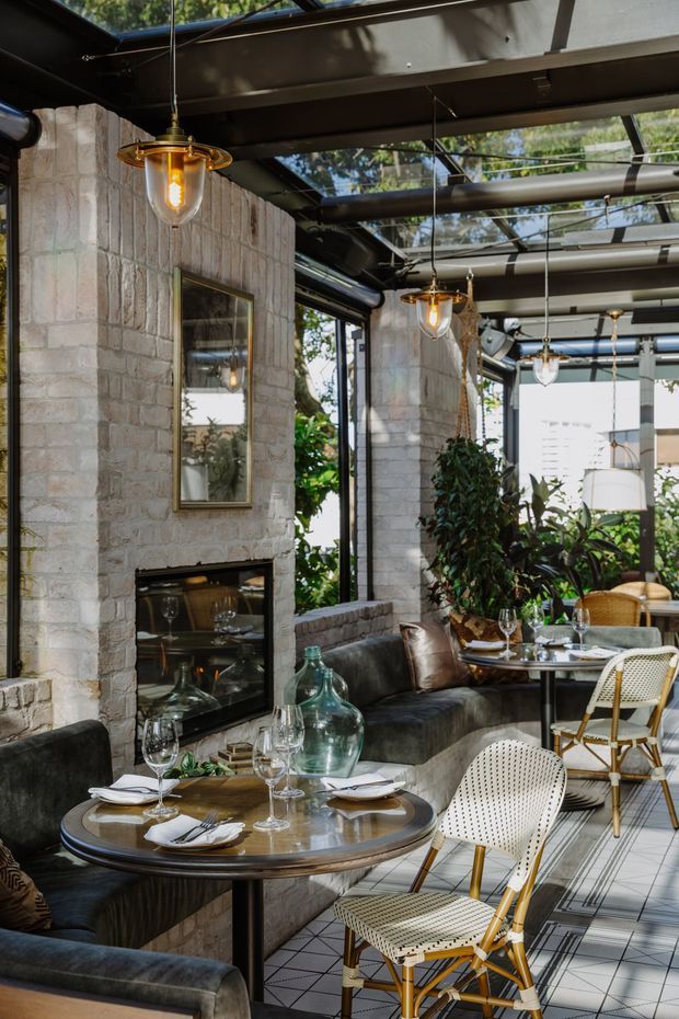 A refined restaurant dining space with round set tables, upholstered banquette seating, woven café chairs, and warm pendant lights. Pale textured masonry walls frame a built-in fireplace, while extensive glass roof panels and glazing bring in abundant daylight and views of greenery. The interior combines dark metal structure, natural stone, dark leather upholstery, timber accents, and patterned floor tiles.