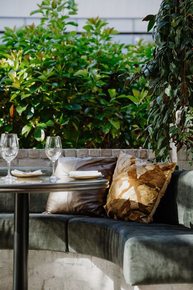 A refined outdoor dining and lounge setting with a curved dark upholstered banquette arranged around a round table, set with plates and wine glasses. Neutral cushions and a patterned accent cushion add warmth, while dense green planting and a low pale masonry wall create a lush courtyard atmosphere.