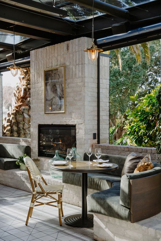 A stylish indoor-outdoor dining and lounge space with a white-painted brick fireplace wall, built-in upholstered banquette seating, a round dining table set with glassware, and a woven café chair. The space has an industrial-style exposed black ceiling with visible ductwork and suspended pendant lights, large open glazed sections, and abundant greenery outside. Geometric tiled flooring and warm natural light create a refined yet relaxed atmosphere.