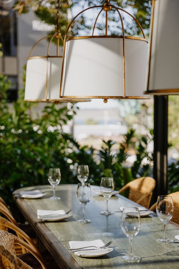 A warmly lit dining area with a long rectangular table set with white napkins, plates, cutlery and multiple wine glasses. Woven rattan dining chairs surround the table, while large white fabric pendant lights with aged brass or bronze frames hang overhead. Lush greenery and daylight visible beyond the glazing create an indoor-outdoor restaurant or hospitality atmosphere.