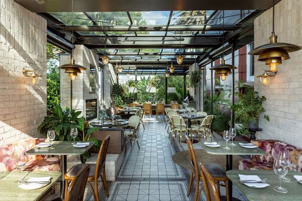 A refined glass-roofed dining space with exposed dark metal framing, pale brick walls, tiled flooring, and abundant greenery. Multiple set dining tables with upholstered and woven chairs line a central aisle, while warm pendant and wall lighting create an inviting restaurant-like atmosphere. The glazed roof and surrounding planting connect the interior to the outdoors.