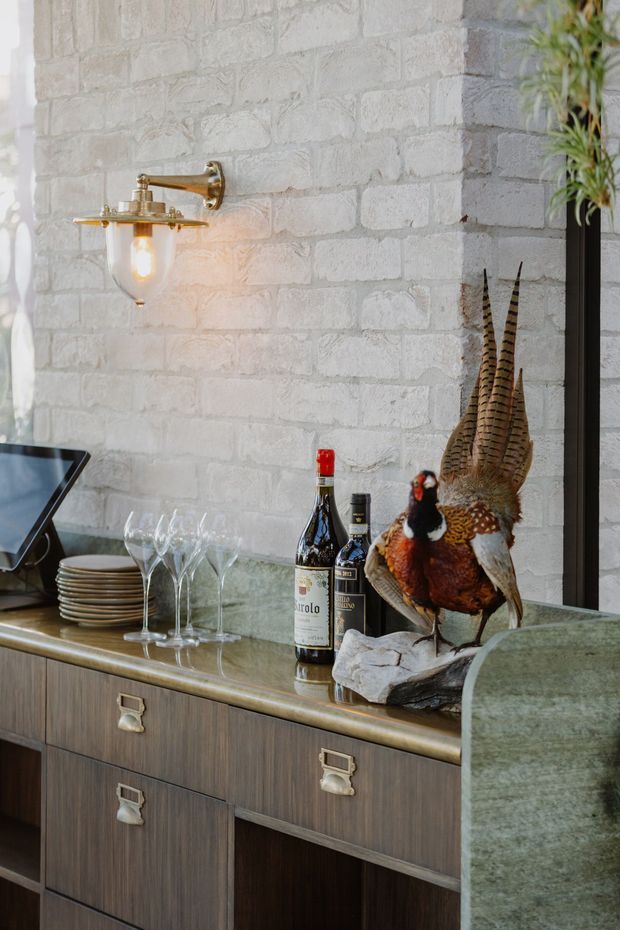 Close-up interior view of a styled home bar or drinks cabinet. A dark timber built-in sideboard with brass-toned hardware supports a greenish stone or glass countertop, wine bottles, stemware and stacked plates. Above it is a brass wall-mounted light with an exposed bulb against a white-painted textured brick wall. A taxidermy pheasant adds a decorative focal point, while a green upholstered or stone-edged partition appears at right.