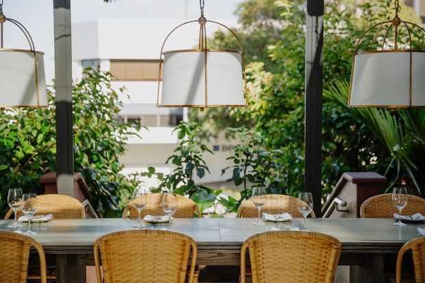 A refined dining area with a long dark dining table set with glassware, napkins and plates, surrounded by woven rattan dining chairs. Large windows overlook lush greenery and a nearby building, while oversized hanging pendant lights with brass frames and white shades provide a warm, elegant focal point. A mirrored wall reflects the dining setting and contributes to the sense of depth.