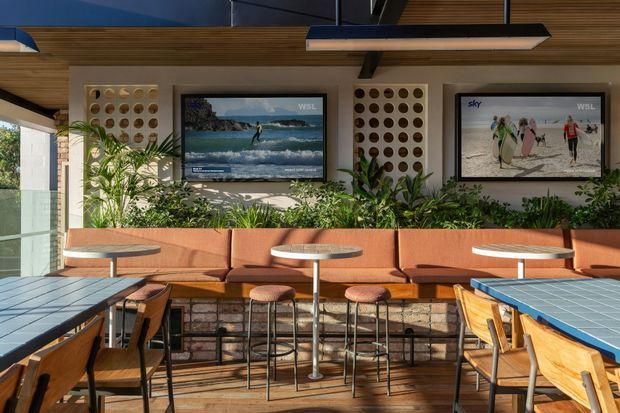 A contemporary hospitality or commercial lounge and dining area with long banquette seating, bar stools, small round tables, and timber dining tables. The space features warm timber ceiling and flooring, exposed dark structural beams, brick and stone lower wall surfaces, blue tiled tabletops, extensive planter greenery, and wall-mounted televisions displaying surfing. Large glazed openings bring in bright natural light.