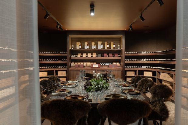 A richly styled private wine room viewed through partially open sheer curtains. Dark timber wine racks line the walls, with bottles, display shelving, and storage drawers surrounding a long dining table set with plates, glassware, candles, and a central leafy arrangement. Fur-covered dining chairs, warm wood tones, and focused track lighting create an intimate, luxurious tasting and entertaining space.