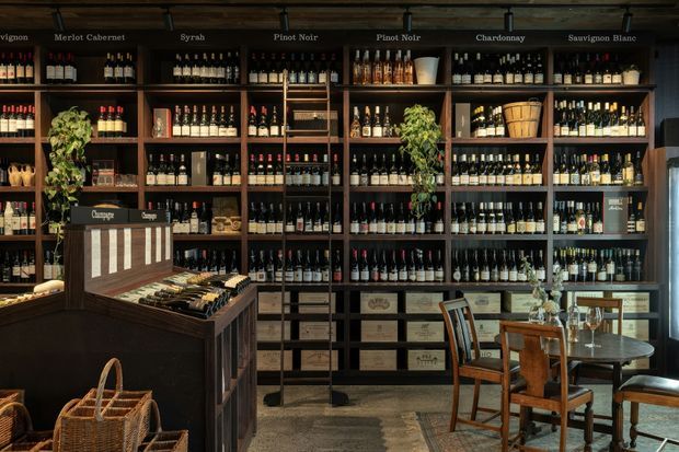 A richly stocked wine shop or wine bar interior with floor-to-ceiling dark timber shelving displaying bottles, labelled wine sections, storage drawers, a central retail counter, and a small dining table with chairs. Warm, low lighting and dark wood create a refined, atmospheric setting.