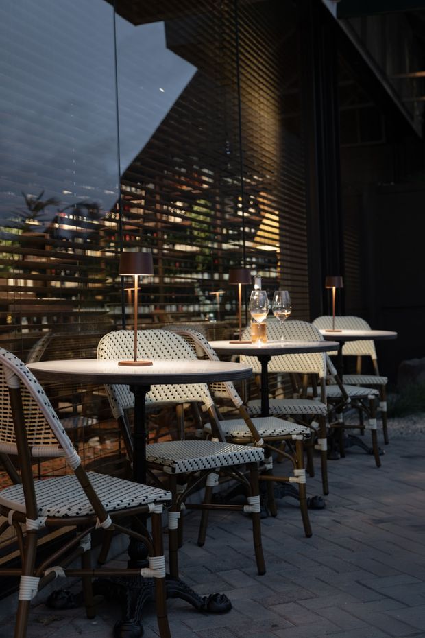 An atmospheric outdoor dining terrace beside a dark glass facade, furnished with small round tables, woven café chairs, table lamps and place settings. Horizontal timber screening and reflections create a layered architectural backdrop, while the paved floor and warm lighting give the space an intimate evening restaurant feel.
