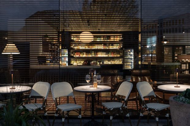 A moody evening view of a sophisticated restaurant or bar interior, seen through horizontal louvres or blinds. Small round tables with candles and wine settings are surrounded by woven lounge chairs, while a warmly illuminated bar and bottle display forms the backdrop. Dark timber and metal finishes, layered lighting, and indoor planting create an intimate hospitality atmosphere.
