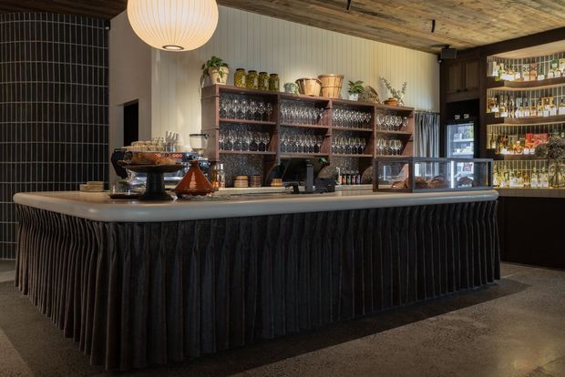 A stylish hospitality bar or café interior with a large central service counter. The counter has a light stone or solid-surface top and a dramatic dark vertically pleated front, paired with dark timber shelving filled with glassware, bottles, and bar accessories. A textured timber ceiling, dark tiled wall, ribbed light wall panelling, warm pendant lighting, and polished concrete flooring create a refined, moody atmosphere.