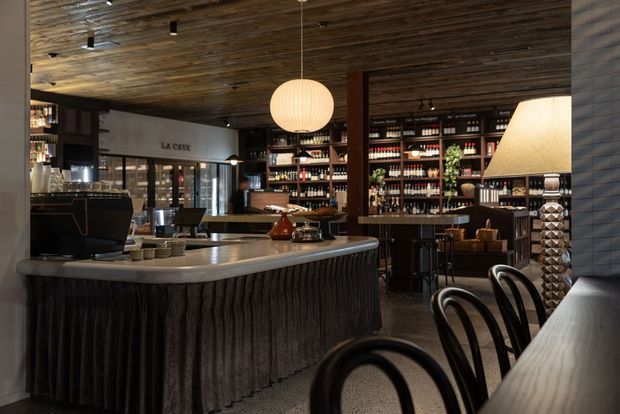 A moody, refined hospitality interior centred on a curved bar or service counter with a pale stone benchtop and pleated dark fabric front. Behind it are extensive timber shelving units displaying bottles, glasses and barware, with a wine cellar labelled “LA CAVE”. Dark timber ceiling boards, warm pendant lighting, table lamps and bentwood bar seating create an intimate restaurant or wine-bar atmosphere.