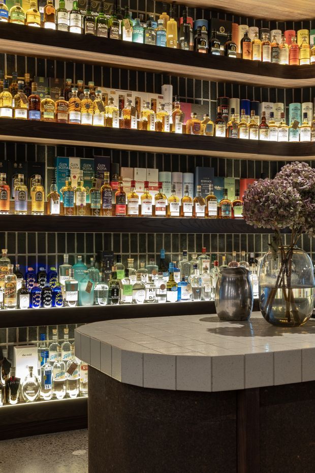 A moody, refined bar interior featuring multiple backlit timber shelves densely stocked with liquor bottles. The shelving is set against a dark vertically gridded tiled wall. In the foreground is a rounded bar island with a pale square-tiled benchtop and dark textured base, accompanied by reflective metalware and a large glass vase containing dried floral foliage.