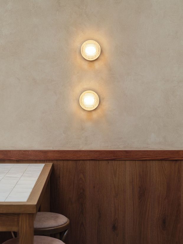 Close-up interior detail showing a warm, textured plaster wall with two vertically aligned round wall sconces. Dark timber wall panelling and trim run along the lower wall, beside a built-in or fixed timber-topped table with a pale tiled surface and a round upholstered stool. The composition is warm, refined, and material-focused.