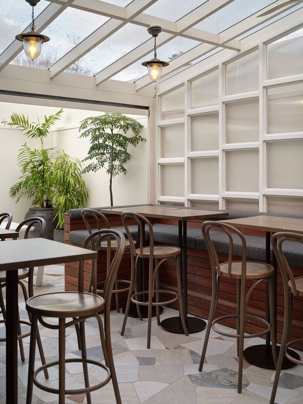 A bright enclosed café dining area with a translucent glazed roof, white-painted structural framing, and a wall of white-framed translucent panels. Dark timber banquette seating and high communal tables are paired with numerous bentwood bar stools. Potted greenery softens the space, while pendant lights hang from the roof structure. The floor is finished in irregular light natural stone tiles with varied grey and beige tones.