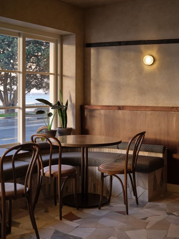A warm, intimate cafe or restaurant dining nook with a round dark-wood table, curved bentwood chairs, and a built-in upholstered banquette. The space features textured earthen plaster walls, dark timber wainscoting and trim, a black horizontal rail, patterned upholstery, irregular natural-stone flooring, and a large divided timber-framed window overlooking a street and greenery. A wall sconce and potted plants add warmth and character.