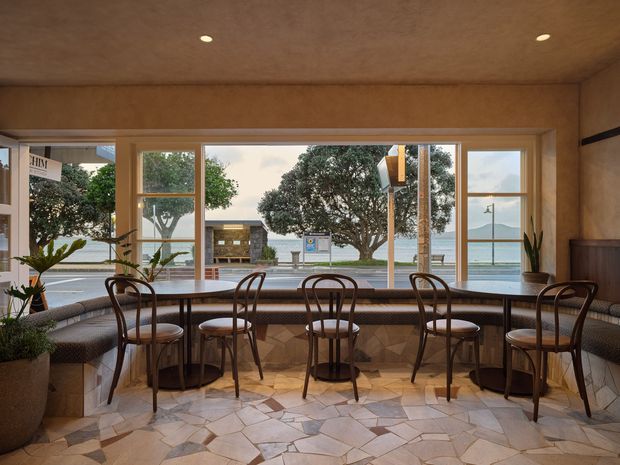 A warm, compact café or hospitality dining space with a long upholstered banquette facing a large glazed window wall, accompanied by several small round tables and bentwood-style chairs. The room features a distinctive irregular stone-tile floor, timber-toned wall and ceiling finishes, recessed downlights, and potted plants. Through the windows, a waterfront promenade and distant hills are visible.