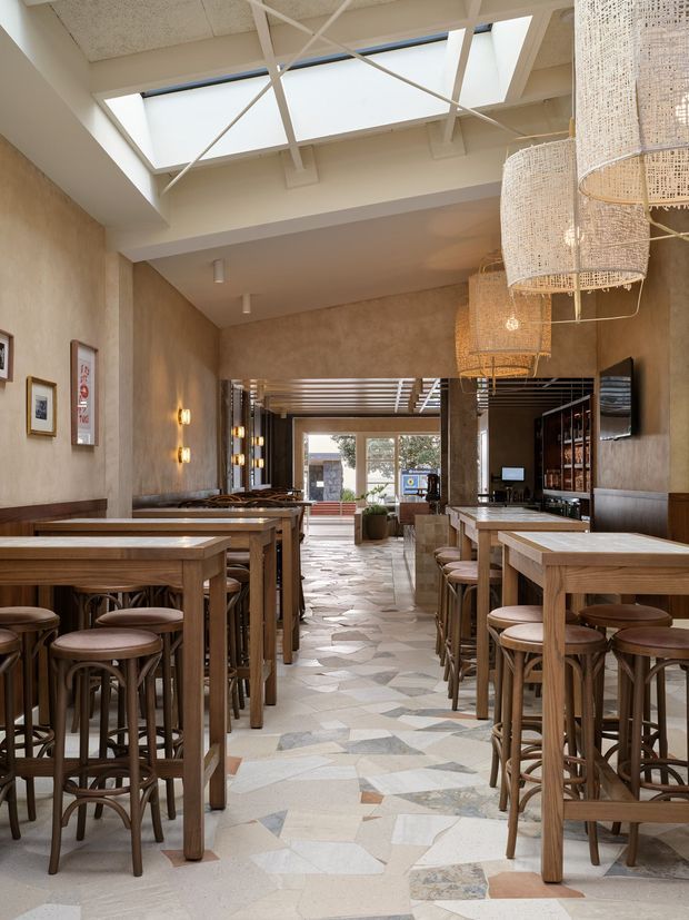 A long, narrow hospitality interior with rows of high timber communal tables and rounded bar stools, textured warm beige walls, a geometric pale stone floor, and a bar or service counter along the right side. Large woven pendant lights hang from a white exposed structural ceiling with skylights, creating a bright, warm, refined atmosphere. The rear opens toward a glazed entrance or window.