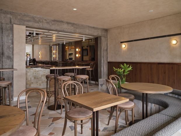 A warmly lit contemporary restaurant or café interior with timber dining tables, bentwood chairs, upholstered banquette seating, a tiled and timber bar, and a concrete-framed opening to the rear dining area. The space combines textured plaster walls, dark timber wall panelling, patterned floor tiles, exposed concrete, and linear wall lighting.