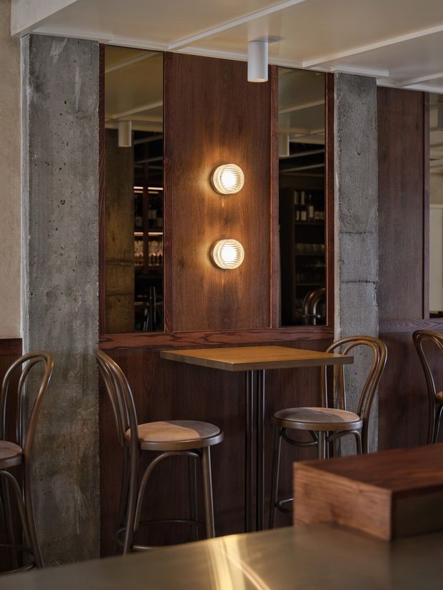 A moody, refined commercial bar or hospitality interior featuring dark timber wall panelling and built-in cabinetry, concrete structural columns, mirrored shelving, warm globe wall lights, and a small timber-topped table with curved bar stools. The space has an industrial-modern character with warm ambient lighting.
