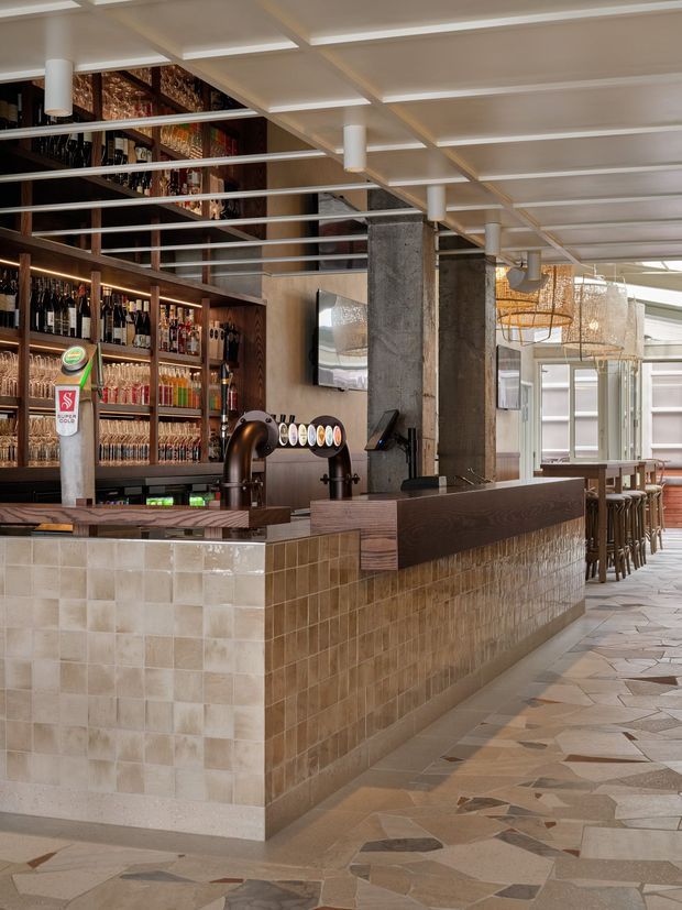 Interior view of a contemporary bar and hospitality venue, featuring a long tiled bar counter, dark timber bottle display and shelving, beer taps, concrete structural columns, upholstered bar stools, and a gridded white ceiling with linear suspended lighting. The floor uses irregular natural stone tiles, and large glazed openings bring in daylight.