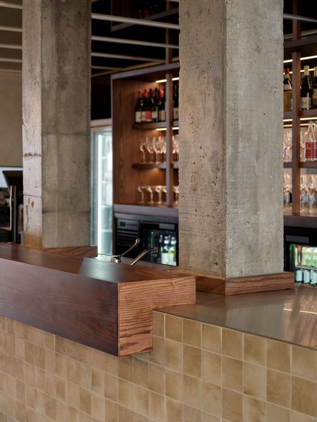 Close-up view of a commercial bar interior with a tiled bar front, dark timber cabinetry and counter detailing, stainless-steel work surface, exposed rough concrete columns, and backlit shelves displaying wine bottles and glassware. The space has a warm, refined industrial character with visible beverage refrigeration and bar fixtures.