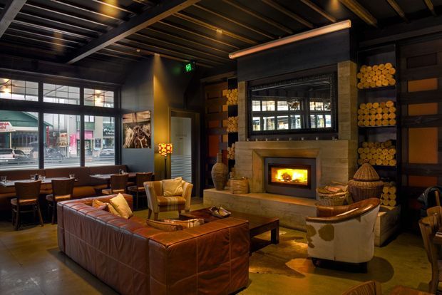 A dark, warmly lit hospitality-style lounge with industrial and rustic detailing. The room features exposed dark ceiling beams, a large storefront window, a central stone fireplace with a glowing fire, built-in shelving stacked with cut logs, leather seating, timber tables, and a cowhide rug. The arrangement creates an intimate bar or restaurant lounge atmosphere.