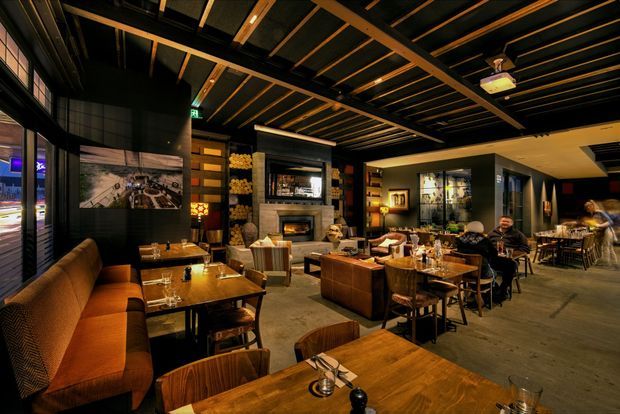 A large, dimly lit restaurant dining interior with warm timber tables, upholstered booth seating, loose dining chairs, and a central fireplace set within a stone surround. The space features exposed dark ceiling beams and timber slats, dark walls, display shelving, artwork, pendant lighting, and extensive glazing along one side. The atmosphere is warm, intimate, and refined, with multiple dining zones extending into the background.