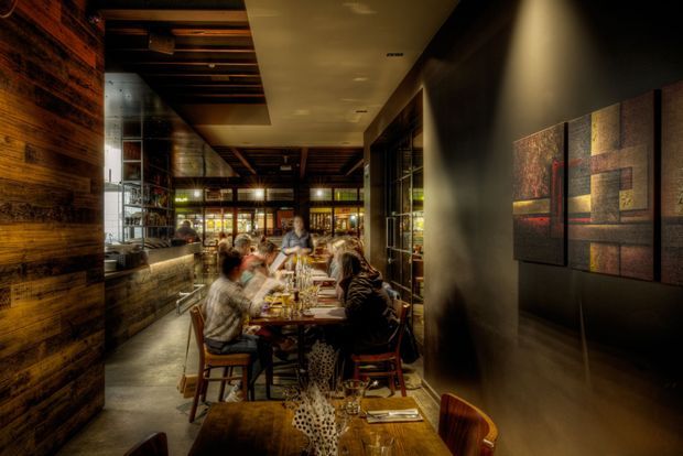 A dimly lit, sophisticated restaurant dining space viewed along a narrow dining room. Guests sit at timber tables with place settings and drinks, surrounded by dark timber wall cladding, exposed timber ceiling beams, glass-fronted shelving, and warm ambient lighting. Abstract geometric artwork decorates the dark wall on the right, creating an intimate, moody atmosphere.