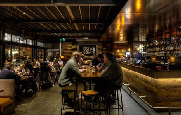 A dimly lit industrial-style restaurant and bar interior, viewed from the dining area. The space features exposed dark ceiling beams, timber and dark metal finishes, a long illuminated timber-fronted bar with extensive bottle shelving, communal and small dining tables, and warm pendant and concealed lighting. Patrons occupy the tables, while large glazed openings line the left side.