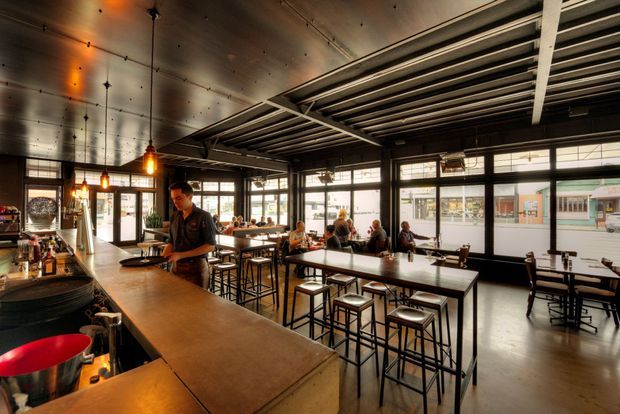 A warm, industrial-style café or bar interior with a long timber service counter, pendant lights, exposed dark ceiling structure, large black-framed windows, and communal high tables with stools. Patrons are seated near the street-facing glazing, while bottles and bar equipment occupy the counter area.