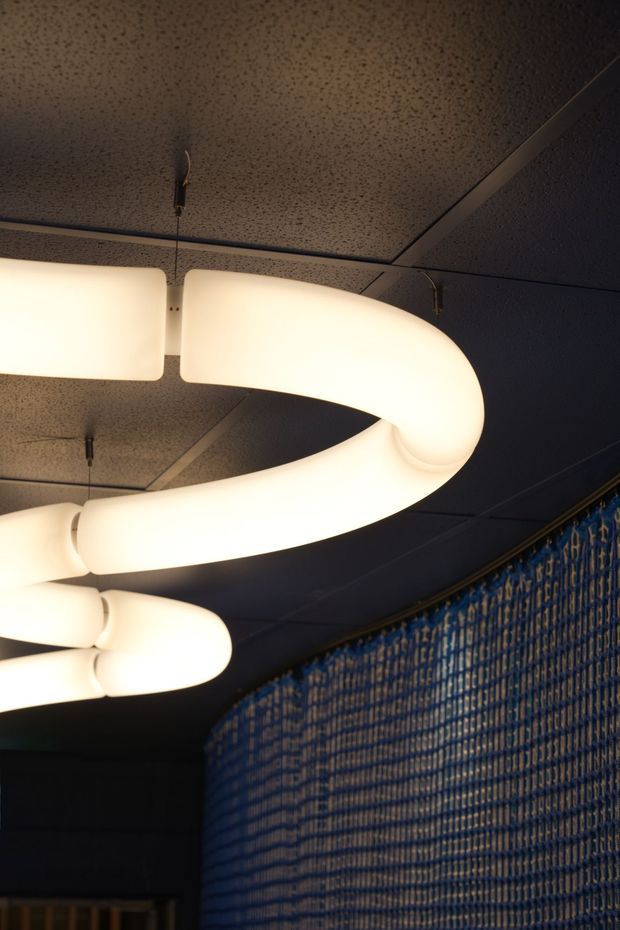 Close-up interior view of a dark, atmospheric space featuring large looping tubular pendant lights with warm white diffused illumination. The ceiling is covered in dark acoustic tiles, while a curved wall is finished with a textured blue patterned surface. The image focuses on dramatic lighting and distinctive interior finishes rather than furniture or room function.